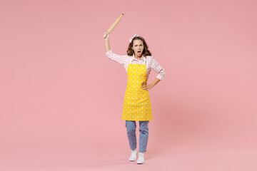 Full length portrait of angry young brunette woman housewife 20s in yellow apron hold rolling pin...