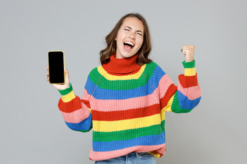 Happy young brunette woman in casual colorful knitted sweater doing winner gesture hold mobile cell phone with blank empty screen mock up copy space isolated on grey colour background studio portrait.