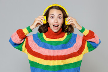 Surprised excited cheerful young brunette woman 20s in casual colorful sweater listening music with headphones keeping mouth open looking camera isolated on grey colour background, studio portrait.
