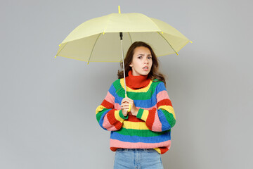 Displeased puzzled concerned shocked worried young woman 20s wearing casual colorful knitted sweater standing under yellow umbrella looking camera isolated on grey colour background, studio portrait.