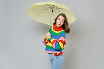 Side view of excited cheerful laughing joyful young woman 20s wearing casual colorful knitted sweater standing under yellow umbrella looking camera isolated on grey colour background, studio portrait.
