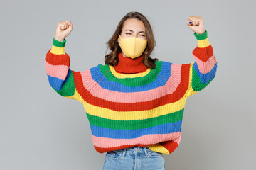 Happy young woman in colorful sweater face mask safe from coronavirus virus covid-19 during pandemic quarantine doing winner gesture keeping eyes closed isolated on grey background, studio portrait.