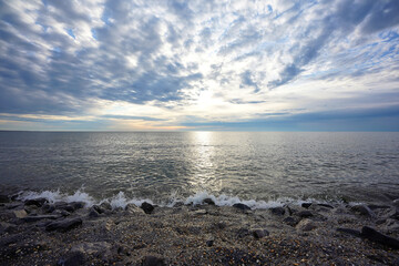 Calm North Sea in the evening, gray stones in the foreground, blue sky with white clouds and yellow sun. Brouwersdam, Netherlands.
