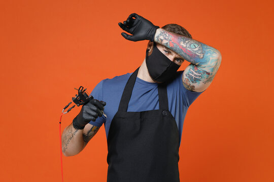 Exhausted Professional Tattooer Master Artist Tattooed Man In T-shirt Apron Face Mask Hold Machine Black Ink Jar Equipment For Making Tattoo Art On Body Put Hand On Head Isolated On Brown Background.