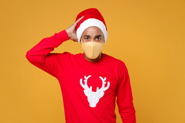 Fototapeta na wymiar Worried young Santa african american man in Christmas hat face mask to safe from coronavirus virus covid-19 put hand on head isolated on yellow background studio. New Year celebration holiday concept.