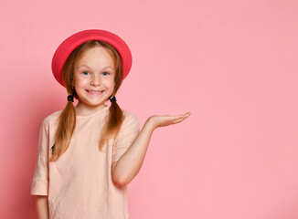 Blonde child in hat and t-shirt. She is smiling and acting like holding something on her palm. Posing on pink studio background. Close up