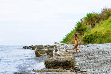 A young girl in a red swimsuit walks along the stones along the seashore with seagulls.