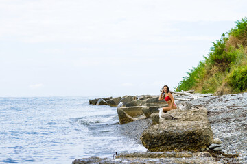 A young girl in a red swimsuit sits on the stones on the seashore with seagulls.