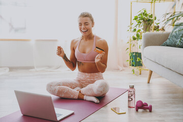 Happy young strong sporty fitness woman wearing pink tracksuit doing yoga exercises using laptop computer hold credit bank card keeping eyes closed sitting and stretching on mat floor at home indoor.