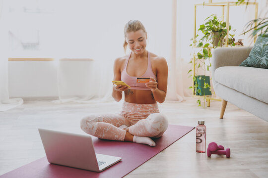 Smiling young strong sporty fitness woman wearing pink tracksuit doing yoga exercises using laptop mobile phone hold credit bank card showing thumb up sitting stretching on mat floor at home indoor.