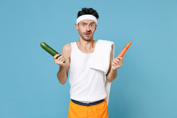 Confused young sporty fitness man with thin skinny body sportsman in white headband shirt shorts hold two vegetables carrot cucumber isolated on blue background. Workout gym sport motivation concept.