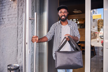 Cheerful young man with delivery bag walking out of cafe
