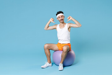 Fototapeta na wymiar Full length portrait excited young man with skinny body sportsman in headband shirt shorts sit on fitball pointing thumbs on himself isolated on blue background. Workout gym sport motivation concept.