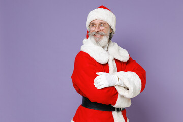 Smiling Santa Claus man in Christmas hat red suit coat white gloves glasses holding hands crossed looking aside isolated on violet background studio. Happy New Year celebration merry holiday concept.
