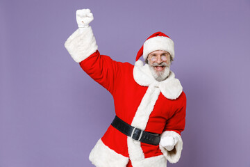 Fototapeta na wymiar Overjoyed Santa Claus man in Christmas hat red coat white gloves glasses clenching fists doing winner gesture isolated on violet background studio. Happy New Year celebration merry holiday concept.