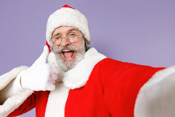 Naklejka premium Close up blinking Santa Claus man in Christmas hat red suit glasses doing selfie shot on mobile phone showing thumb up isolated on violet background. Happy New Year celebration merry holiday concept.