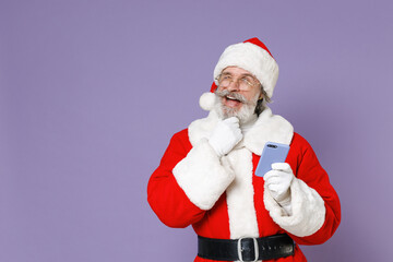 Fototapeta na wymiar Pensive Santa Claus man in Christmas hat red suit coat white gloves glasses using mobile phone put hand prop up on chin isolated on violet background. Happy New Year celebration merry holiday concept.