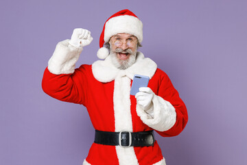 Fototapeta na wymiar Joyful Santa Claus man in Christmas hat red suit coat white gloves glasses using mobile phone doing winner gesture isolated on violet background. Happy New Year celebration merry holiday concept.