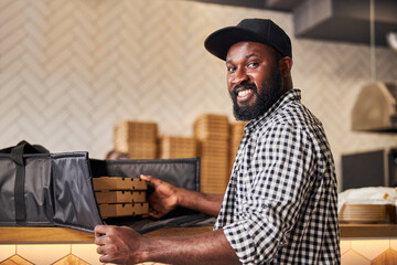 Joyful Afro American man putting pizza boxes in delivery bag