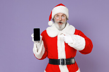 Shocked Santa Claus man in Christmas hat red coat glasses pointing index finger on mobile phone with blank empty screen isolated on violet background. Happy New Year celebration merry holiday concept.