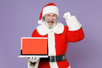 Joyful Santa Claus man in Christmas hat red coat hold laptop pc computer with blank empty screen doing winner gesture isolated on violet background. Happy New Year celebration merry holiday concept.