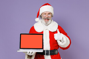 Blinking Santa Claus man in Christmas hat red coat hold laptop pc computer with blank empty screen showing thumb up isolated on violet background. Happy New Year celebration merry holiday concept.