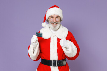 Fototapeta na wymiar Smiling elderly Santa Claus man in Christmas hat red suit coat gloves glasses hold credit bank card showing thumb up isolated on violet background. Happy New Year celebration merry holiday concept.
