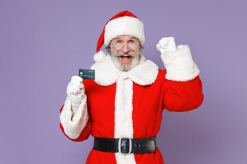 Fototapeta na wymiar Joyful elderly Santa Claus man in Christmas hat red suit coat gloves glasses hold credit bank card doing winner gesture isolated on violet background. Happy New Year celebration merry holiday concept.