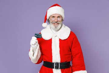 Smiling elderly gray-haired Santa Claus man in Christmas hat red suit coat white gloves glasses hold credit bank card isolated on violet background. Happy New Year celebration merry holiday concept.