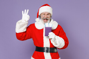 Fototapeta na wymiar Blinking traveler tourist Santa Claus man in Christmas hat red suit glasses hold passport tickets showing OK gesture isolated on violet background. Happy New Year celebration merry holiday concept.