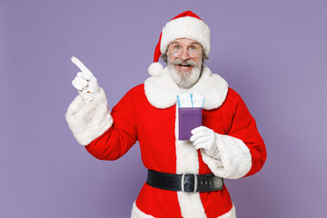 Fototapeta na wymiar Amazed traveler tourist Santa Claus man in Christmas hat suit glasses hold passport tickets pointing index finger aside isolated on violet background. Happy New Year celebration merry holiday concept.