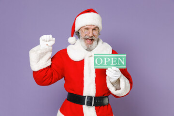 Fototapeta na wymiar Joyful Santa Claus man in Christmas hat red suit coat gloves glasses hold sign with OPEN title doing winner gesture isolated on violet background. Happy New Year celebration merry holiday concept.