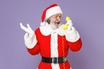 Excited Santa Claus man in Christmas hat red suit coat white gloves glasses talking on telephone spreading hands isolated on violet background studio. Happy New Year celebration merry holiday concept.