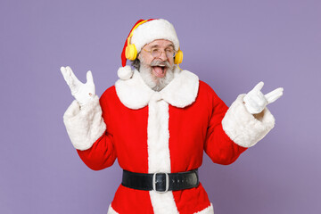 Fototapeta na wymiar Cheerful Santa Claus man in Christmas hat red suit coat gloves glasses listening music with headphones spreading hands isolated on violet background. Happy New Year celebration merry holiday concept.