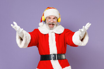 Fototapeta na wymiar Laughing Santa Claus man in Christmas hat red suit coat gloves glasses listening music with headphones spreading hands isolated on violet background. Happy New Year celebration merry holiday concept.