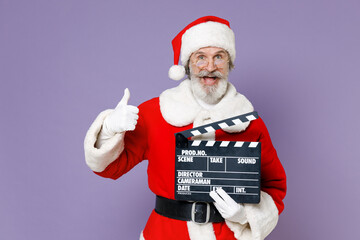 Excited Santa Claus man in Christmas hat red coat glasses showing thumb up hold classic black film making clapperboard isolated on violet background. Happy New Year celebration merry holiday concept.