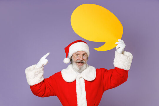 Excited Santa Claus man in Christmas hat red coat gloves glasses pointing index finger on yellow empty blank Say cloud isolated on violet background. Happy New Year celebration merry holiday concept.