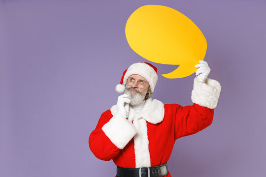 Pensive Santa Claus man in Christmas hat red coat glasses put hand prop up on chin hold yellow blank empty Say cloud isolated on violet background. Happy New Year celebration merry holiday concept.