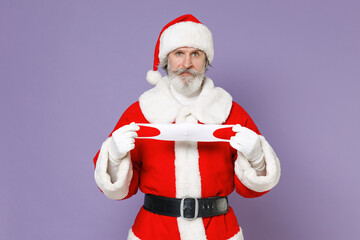 Displeased Santa Claus man in Christmas hat red coat hold face mask to safe from coronavirus virus covid-19 during quarantine isolated on violet background. New Year celebration merry holiday concept.