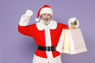 Joyful Santa Claus man in Christmas hat coat glasses hold package bags purchases after shopping doing winner gesture isolated on violet background. Happy New Year celebration merry holiday concept.