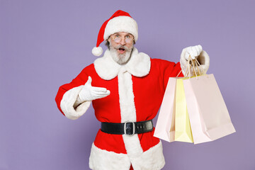 Shocked Santa Claus man in Christmas hat red coat suit glasses pointing hand on package bags purchases after shopping isolated on violet background. Happy New Year celebration merry holiday concept.