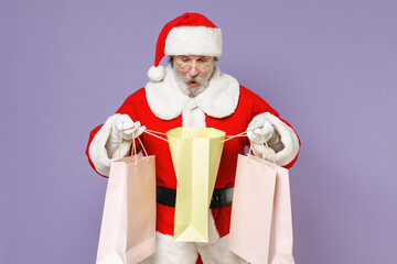 Shocked Santa Claus man in Christmas hat red coat suit white gloves glasses hold package bags purchases after shopping isolated on violet background. Happy New Year celebration merry holiday concept.