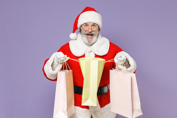 Excited Santa Claus man in Christmas hat red coat suit white gloves glasses hold package bags purchases after shopping isolated on violet background. Happy New Year celebration merry holiday concept.