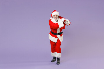 Full length portrait of cheerful Santa Claus man in Christmas hat red coat suit glasses hold bag with presents gifts isolated on violet background. Happy New Year celebration merry holiday concept.