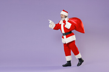 Full length side view portrait amazed Santa Claus man in Christmas hat coat hold bag with presents gifts point finger aside isolated on violet background. Happy New Year celebration holiday concept.