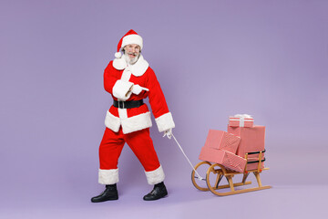 Full length portrait of amazed Santa Claus man in Christmas hat suit pointing index finger on sleigh with present gifts boxes isolated on violet background. Happy New Year celebration holiday concept.