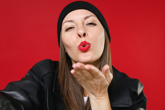 Close up of pretty beautiful young brunette woman 20s wearing casual black leather jacket hat doing selfie shot on mobile phone blowing sending air kiss isolated on red background studio portrait.