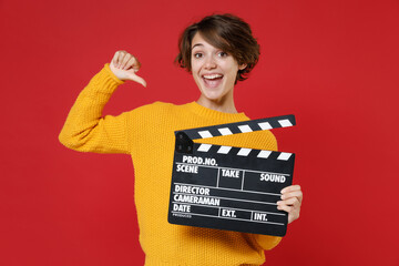 Cheerful young brunette woman 20s in casual yellow sweater standing holding classic black film making clapperboard pointing thumb on herself isolated on bright red colour background studio portrait.