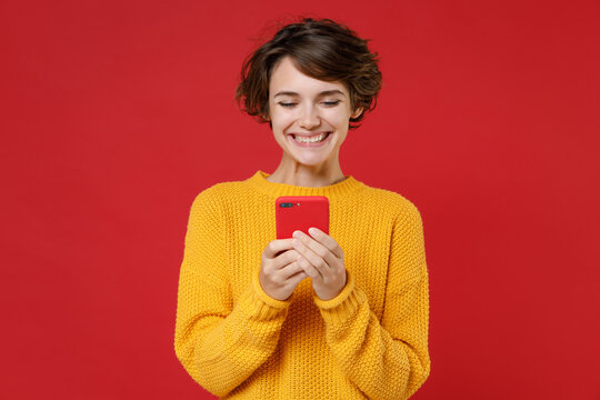 Smiling cheerful pretty attractive young brunette woman 20s wearing casual yellow sweater standing using mobile cell phone typing sms message isolated on bright red colour background studio portrait.