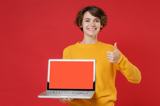 Smiling young brunette woman 20s wearing yellow sweater standing hold laptop pc computer with blank empty screen mock up copy space showing thumb up isolated on bright red background studio portrait.